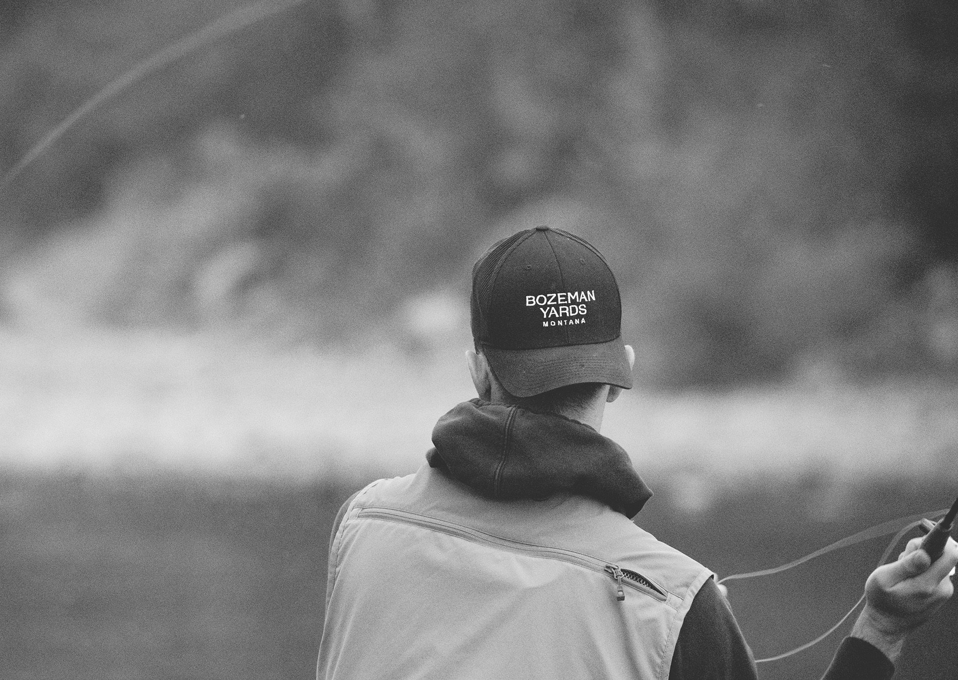 black and white image from behind of a man wearing a Bozeman Yards baseball cap backwards about to cast a fishing rod